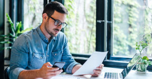 A small business owner reviews paperwork at a desk with a laptop and smartphone.